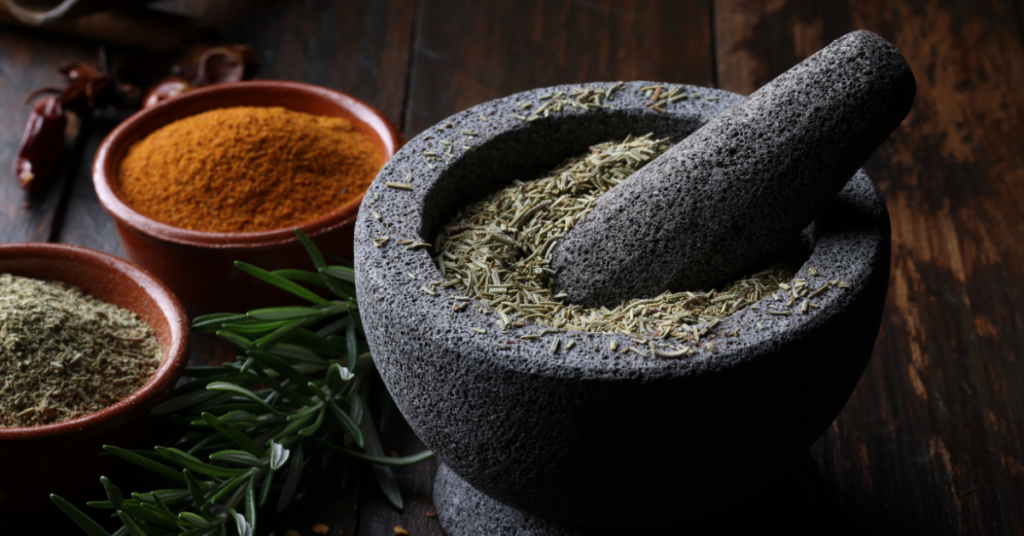 Granite mortar and pestle filled with dried rosemary, surrounded by fresh herbs and spices like turmeric and chili powder, emphasizing the art of creating herbal health remedies.